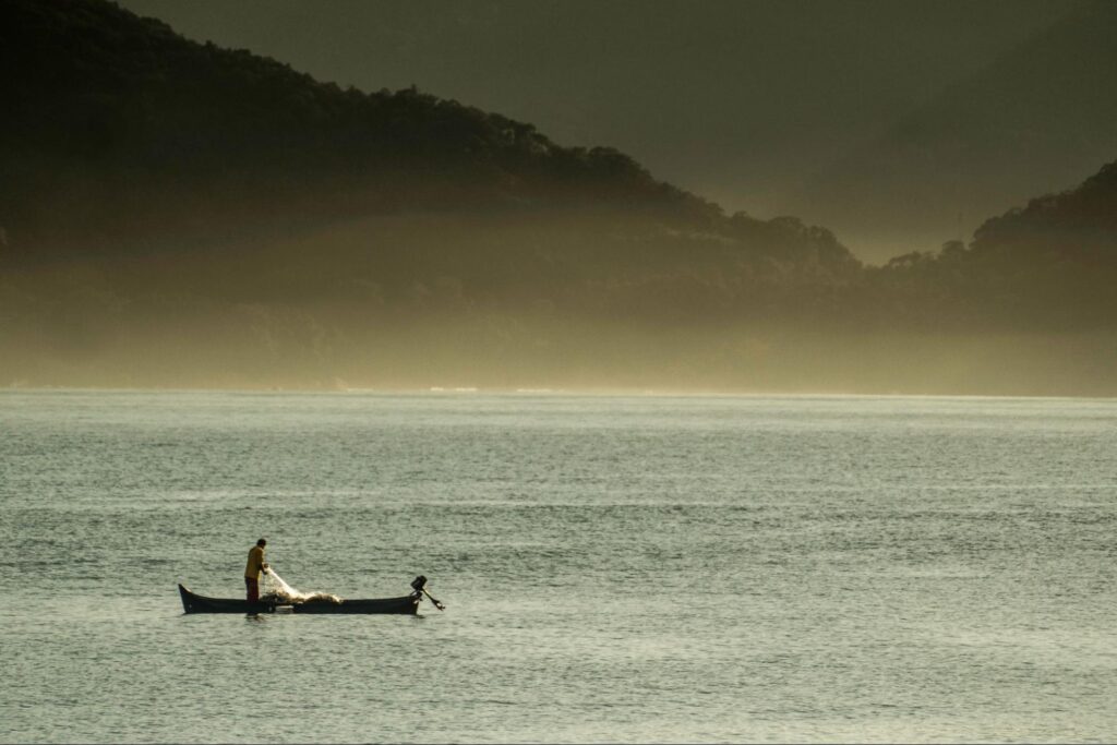 Pescador caiçara no mar, a bordo de uma canoa. Ele se prepara para jogar uma rede de pesca na água. Em segundo plano, aparecem montanhas cobertas pela mata.