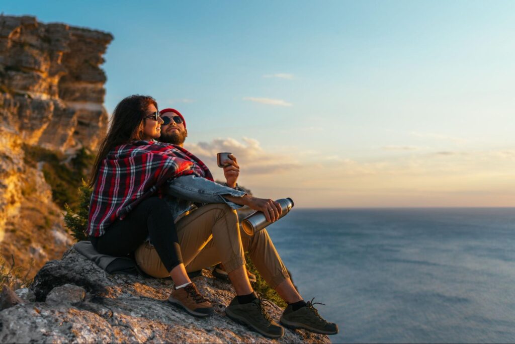 Casal composto por duas mulheres jovens caminhando pela areia de uma praia. Elas se abraçam lateralmente e sorriem uma para a outra.