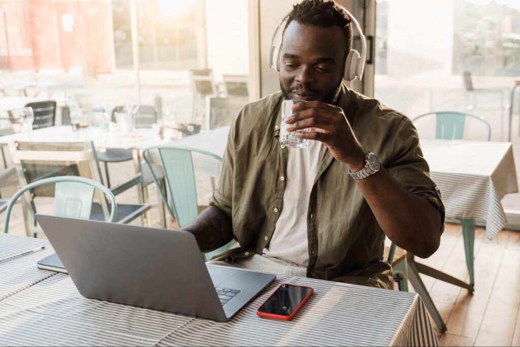 (Descrição) Homem sentado à mesa em uma cafeteria durante o dia. Ele usa fones de ouvido tipo headphones e olha para a tela do notebook que está aberto à sua frente.