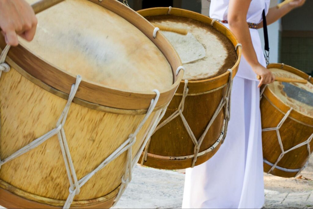 Trio de músicos portando alfaias, grandes tambores feitos de madeira e corda usados tradicionalmente no Maracatu.