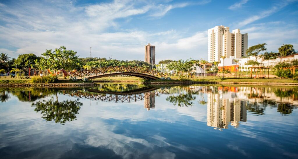 Lago do Parque Ipiranga refletindo o céu azul com nuvens brancas. Área verde com árvores e uma ponte sob o lago.