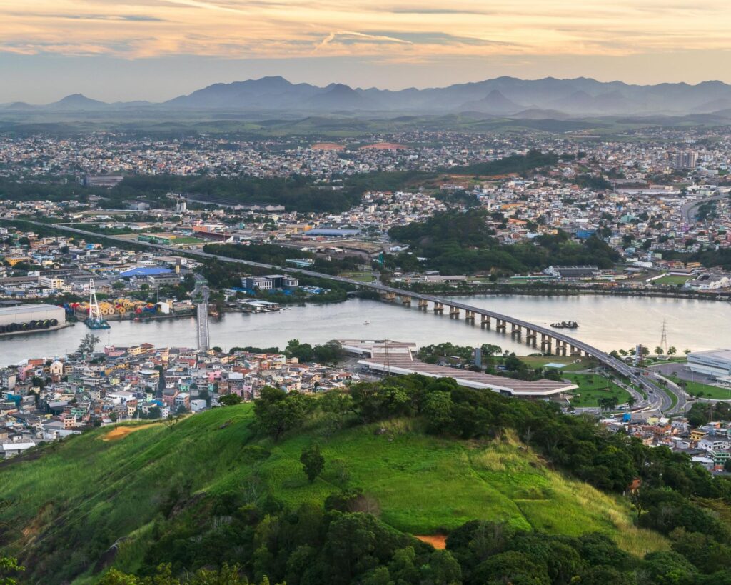 Vista aérea da ponte que liga a cidade de Vitória a Cariacica. Inúmeras casas e prédios em ambos os sentidos, com área verde montanhosa.