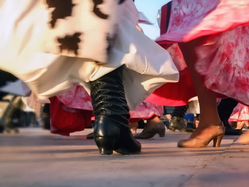 Pés dos dançarinos durante uma coreografia de fandango. Os homens usam botas de meio cano pretas e as mulheres usam sapatos com saltos baixos. As saias rodadas das dançarinas são estampadas em vermelho e branco.