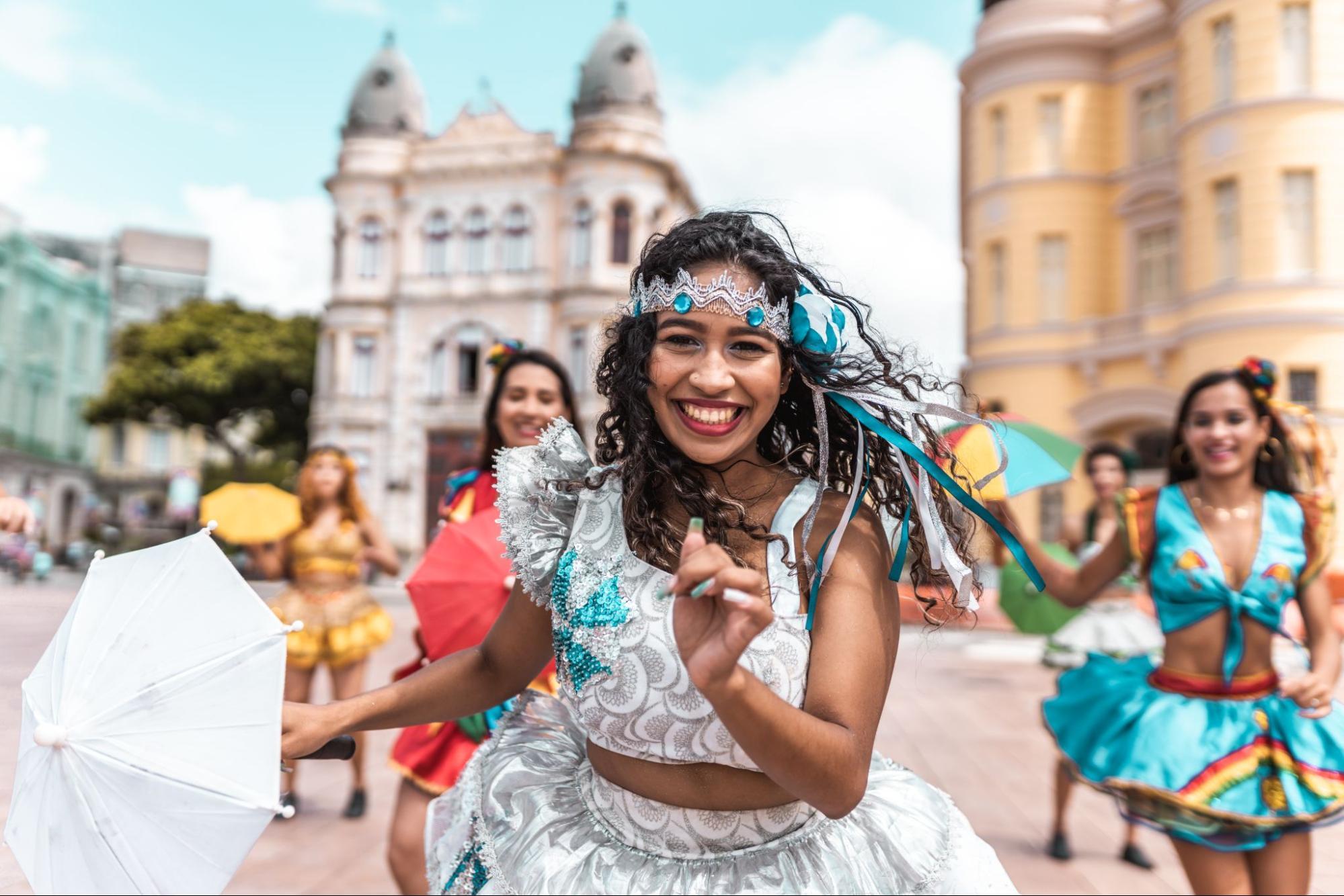 Grupo de passistas dançando frevo no Recife Antigo. Elas usam trajes coloridos bordados com lantejoulas e seguram as tradicionais sombrinhas de frevo.