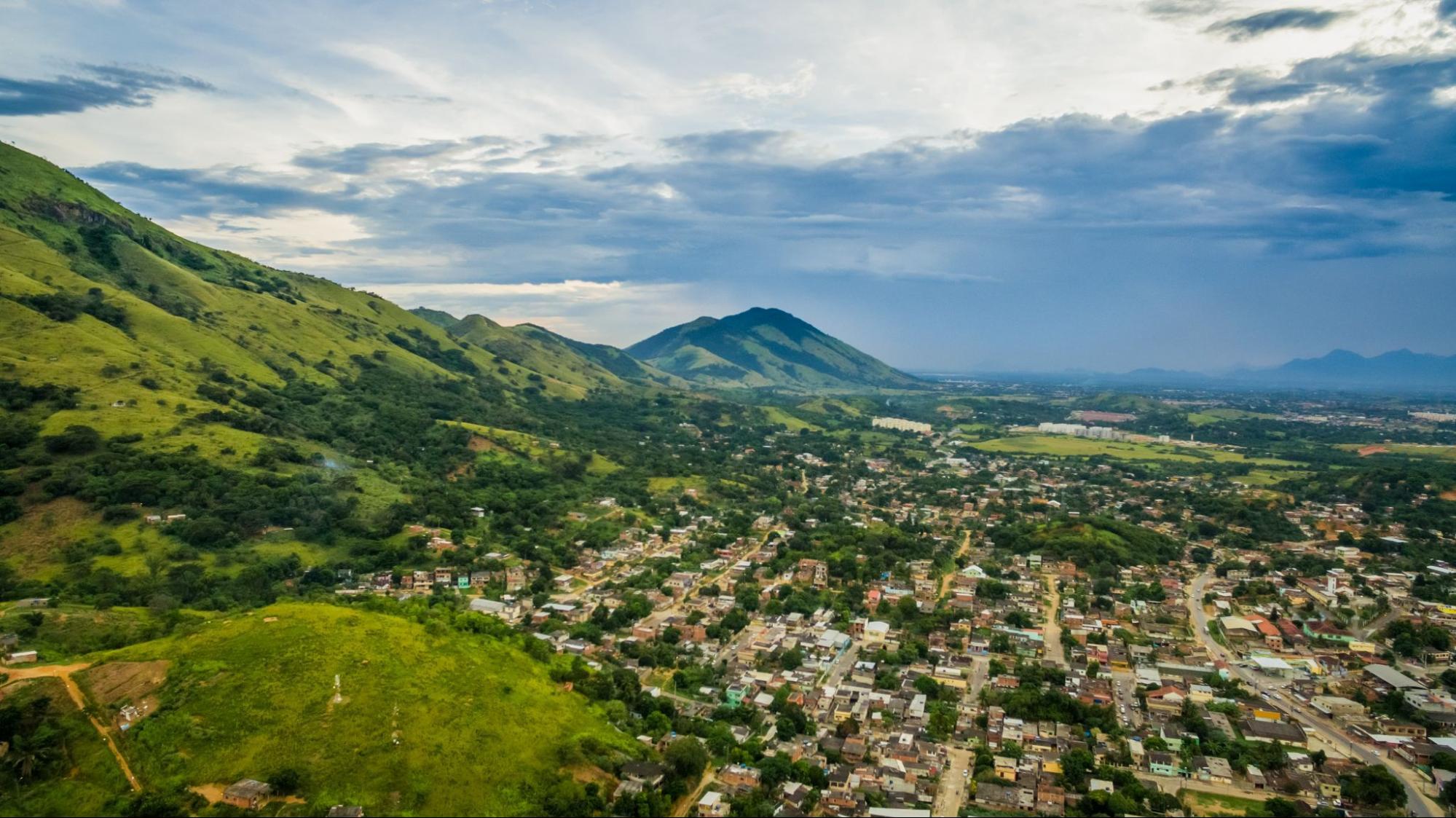 Imagem panorâmica com grandes montanhas verdes ao lado esquerdo, cercando a cidade que se alonga ao canto direito da imagem.
