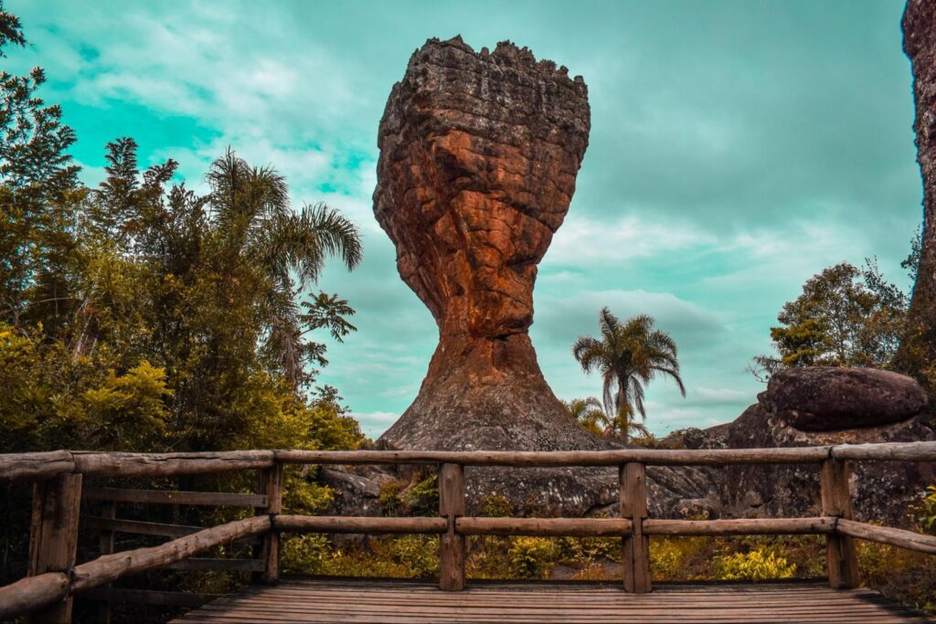 Monumento rochoso naturalmente esculpido no formato de uma taça, com árvores de diferentes espécies ao seu redor e o deck com apoios de madeira do mirante para a vista no Parque Vila Velha.