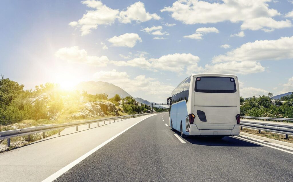 Onibus branco andando em direção à estrada, num céu azul com poucas nuvens e vegetação ao lado.